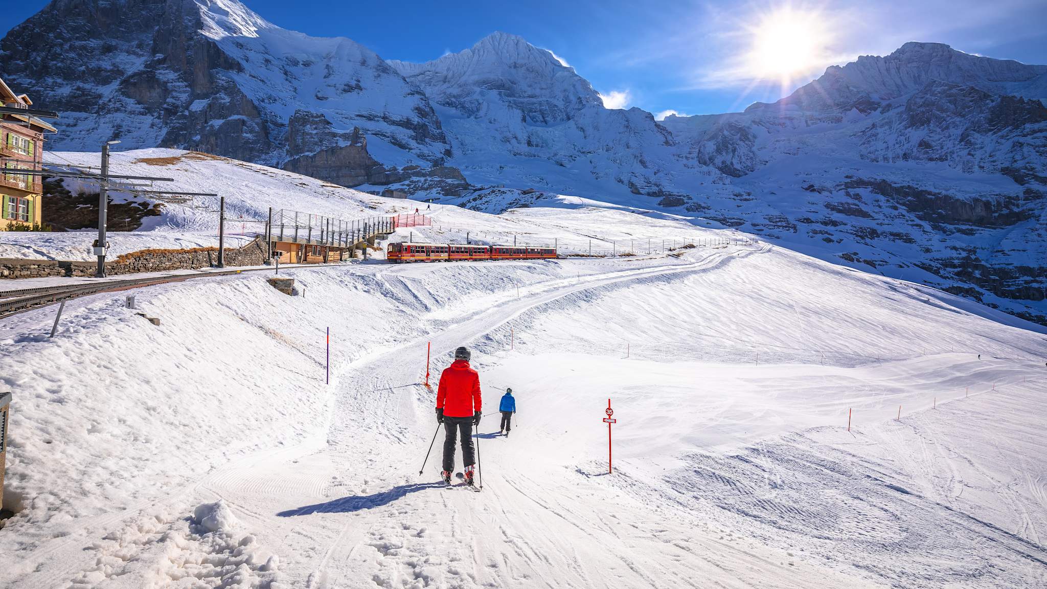 Eine Bahn fährt durch eine verschneite Berglandschaft. Daneben fahren zwei Skifahrer den Berg hinab.