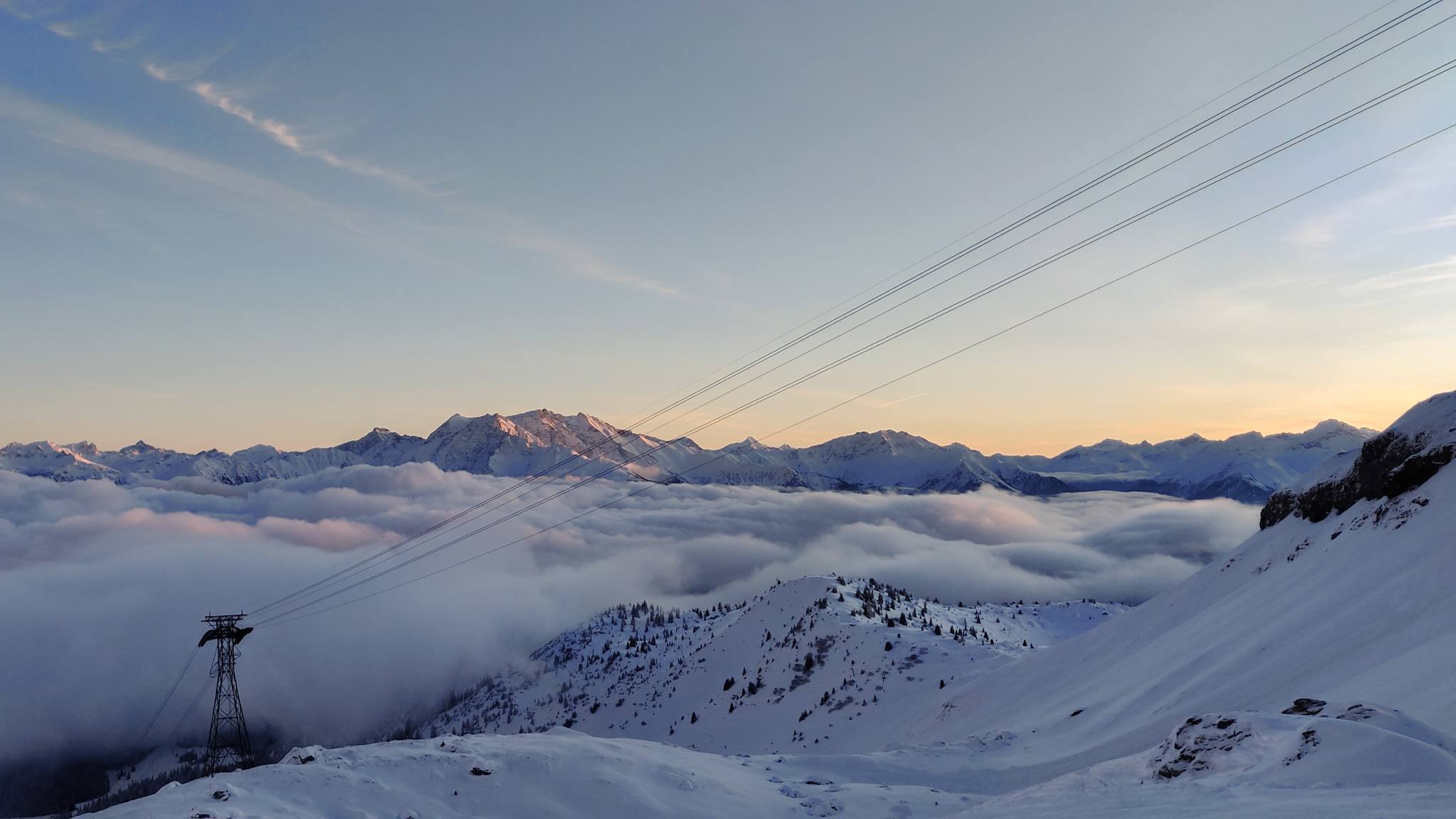 Berge ragen bei Sonnenuntergang aus einem Wolkenmeer heraus. Im Vordergrund verlaufen die Seile einer Seilbahn.