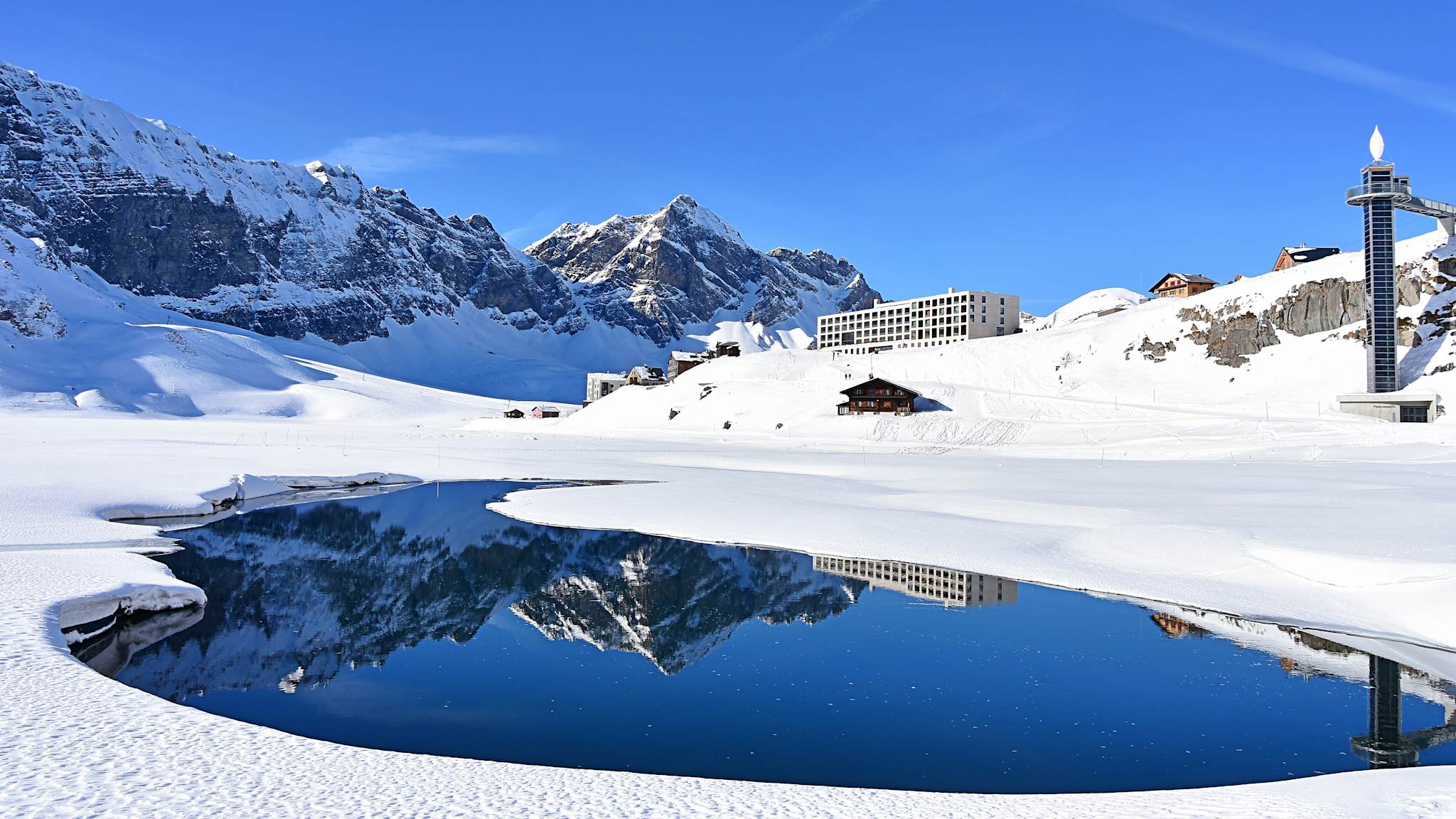 Ein Gebäude und Berge spiegeln sich in einem Bergsee in einer verschneiten Landschaft.