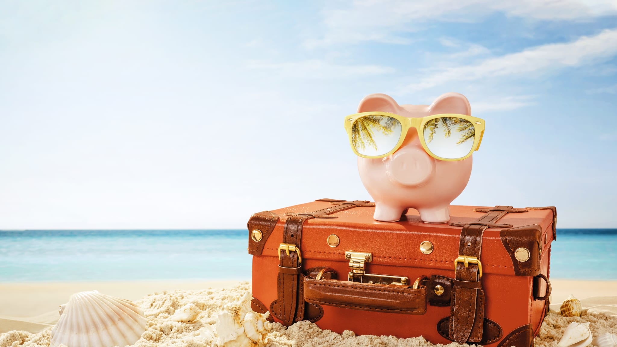 Sparschwein mit Sonnenbrille und Reisekoffer am Strand