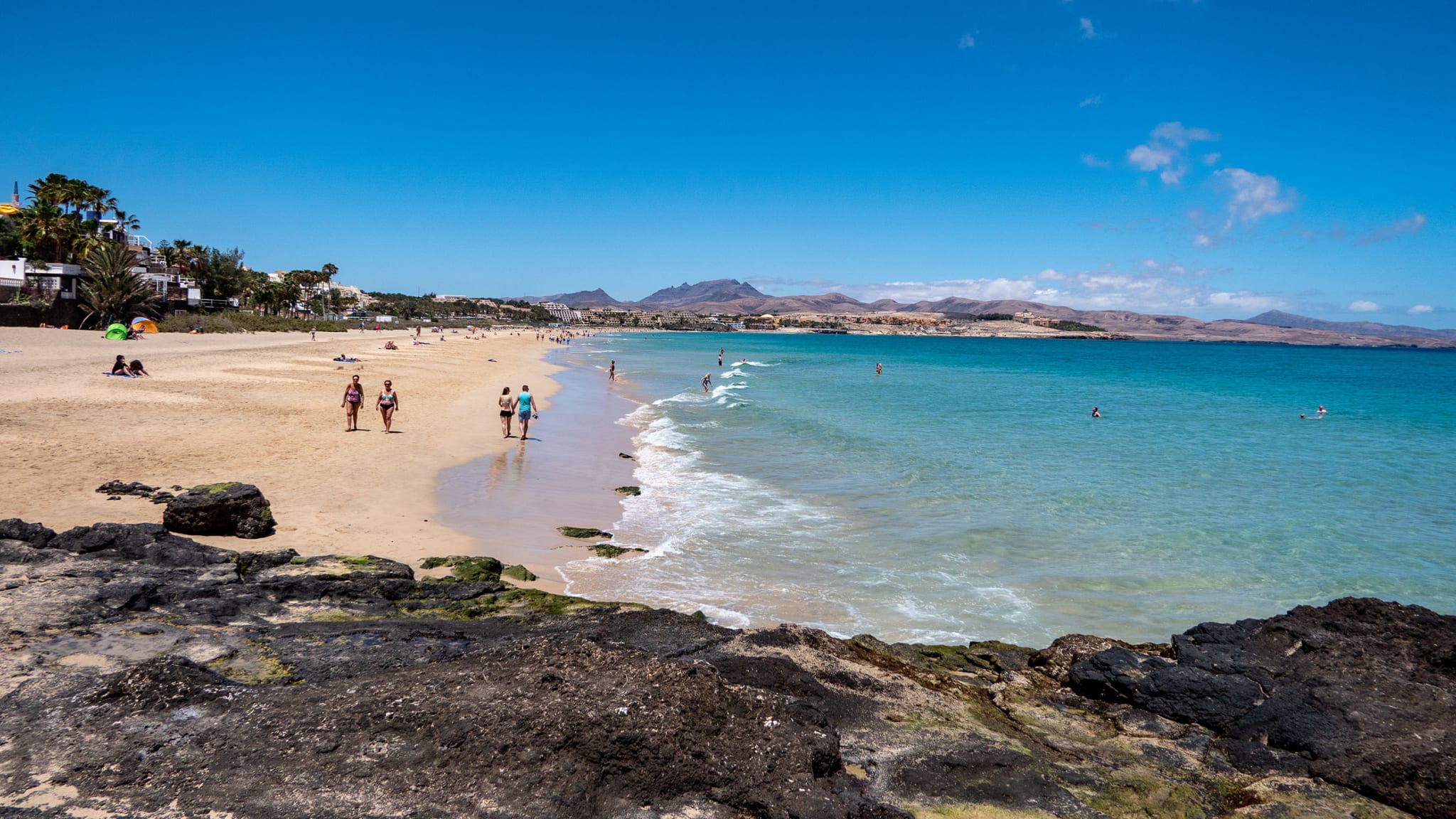 Langer Sandstrand mit Spaziergängern, klares blaues Meer, an der grünen Promenade reihen sich Häuser aneinander auf Fuerteventura