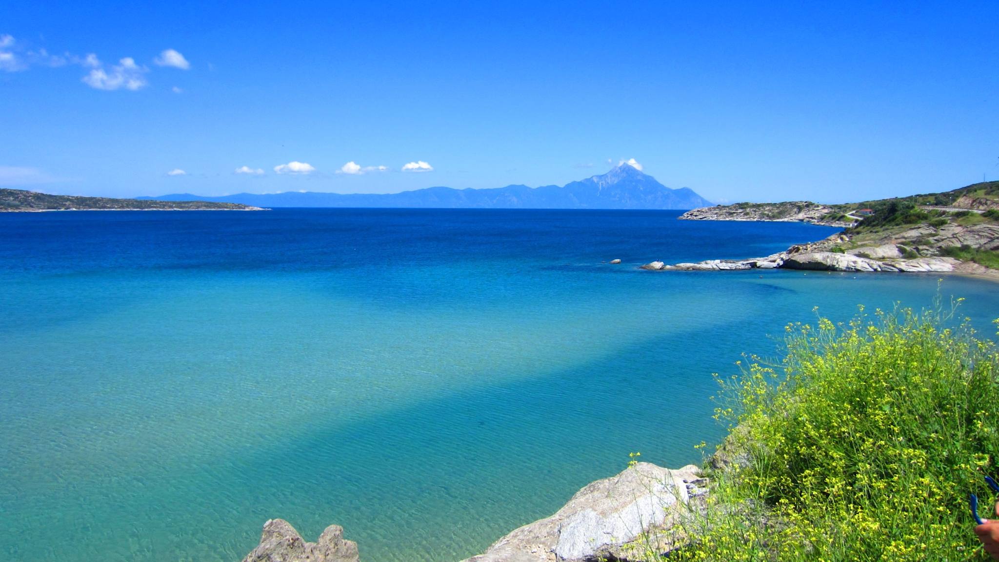 Blick über eine weite Bucht mit blauem Wasser und Bergen im Hintergrund.