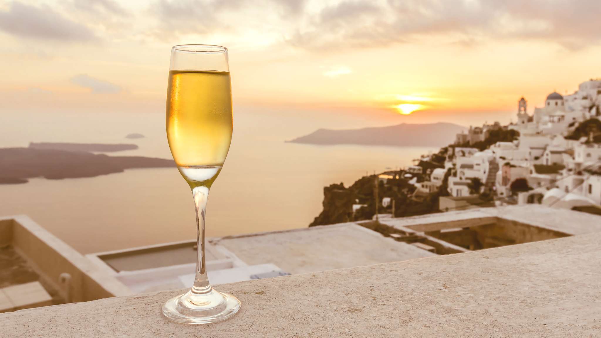 Ein Glas Weißwein steht auf einer Mauer, im Hintergrund sieht man die Caldera Santorinis und die untergehende Sonne