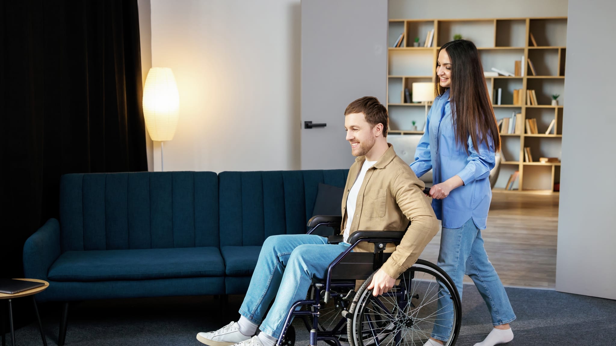 Eine Frau schiebt einen Mann im Rollstuhl durch ein Zimmer.