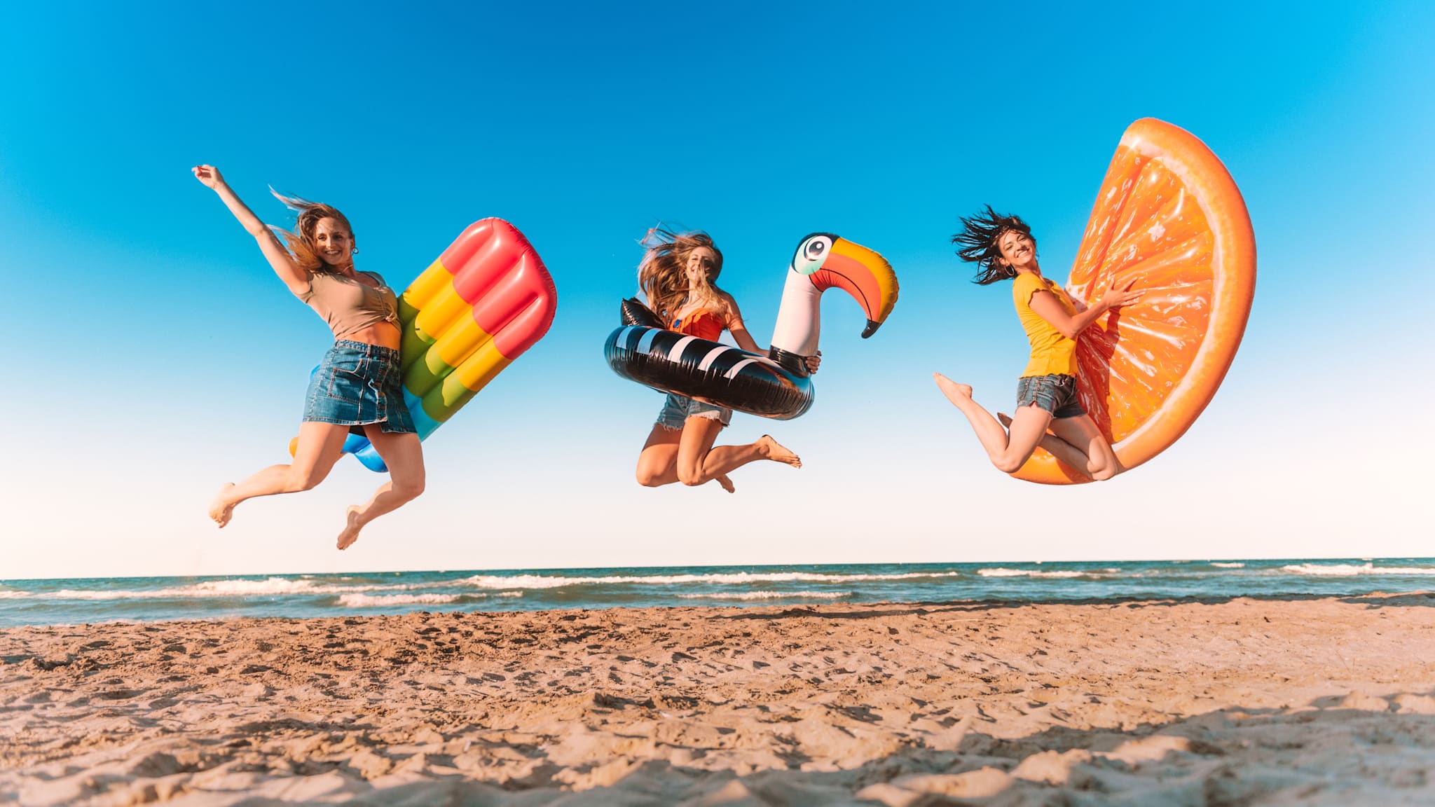 Drei Freundinnen springen mit aufblasbarem Spielzeug am Strand.