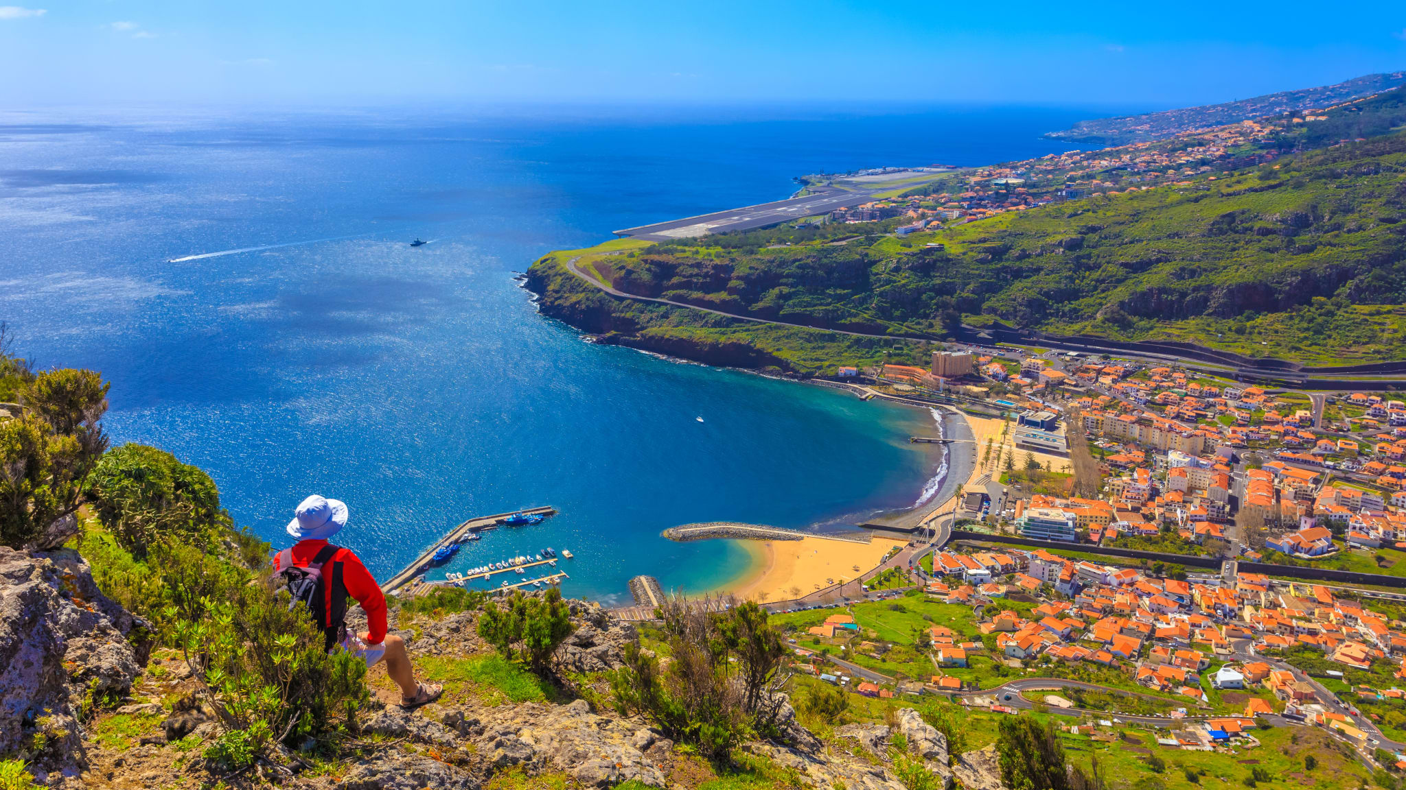 Im Vordergrund sitzt eine Person und blickt über die Machico Bucht zum Flughafen von Madeira.