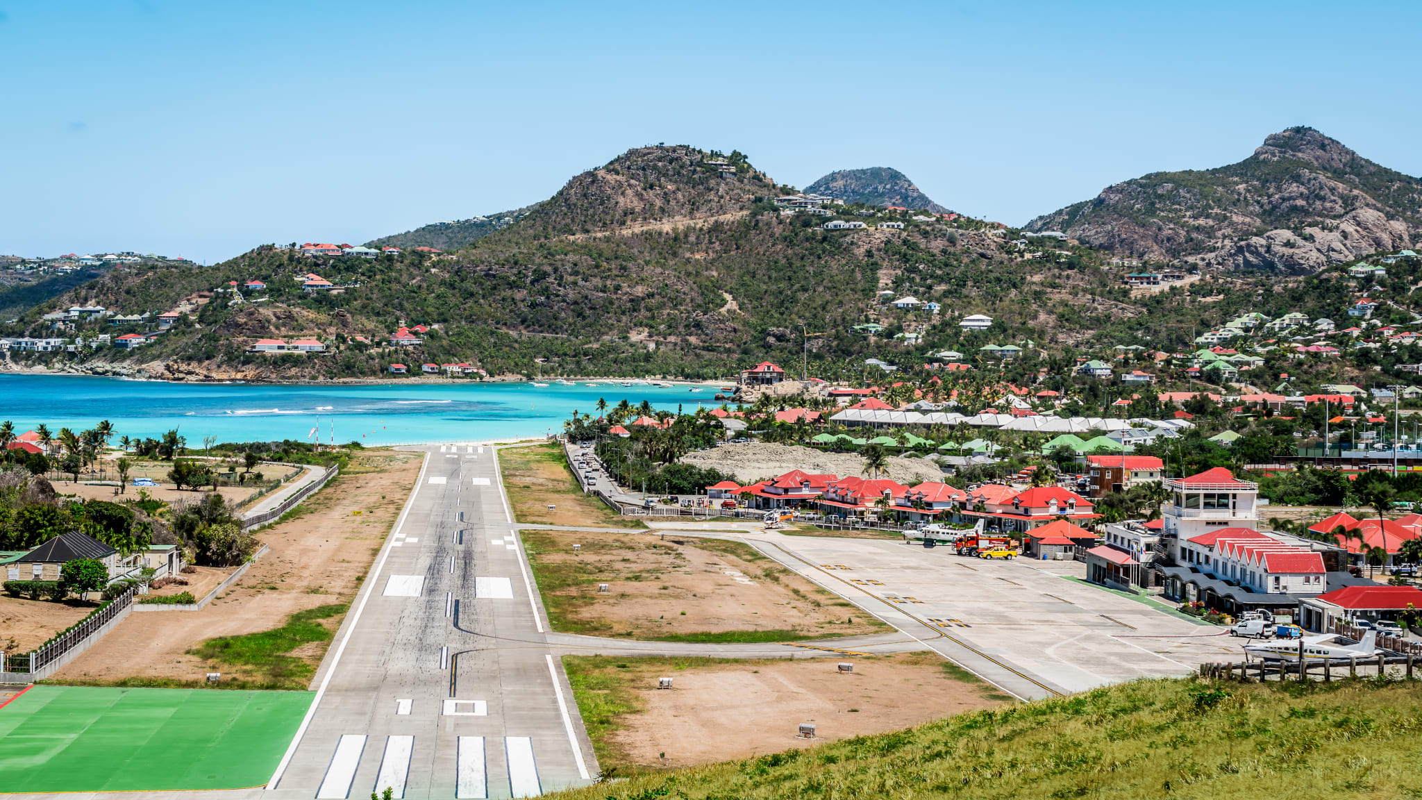 Im Vordergrund ist der kurze Runway von St.Barth zu sehen, dahinter eine Bucht mit Strand und Bergen.