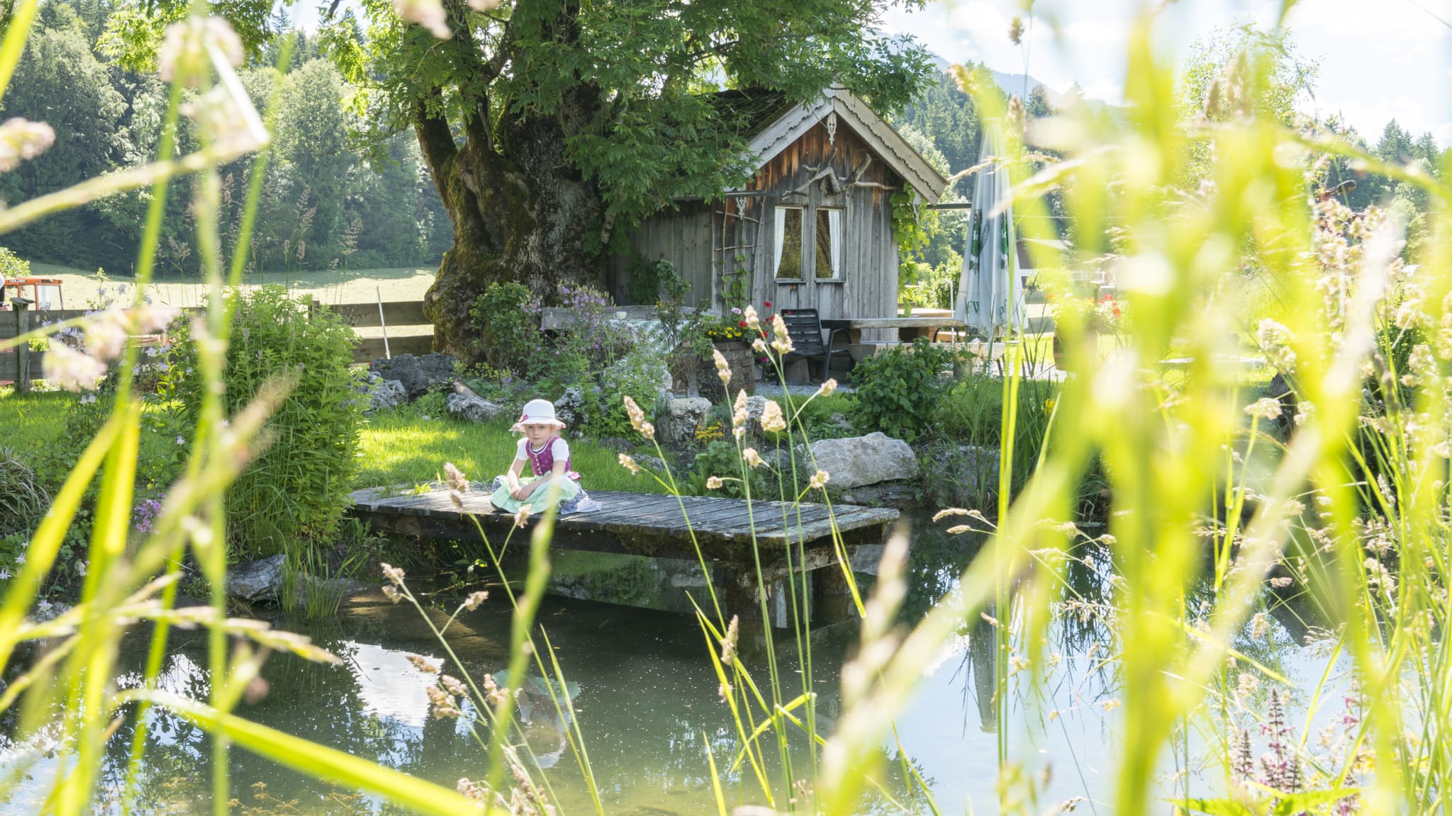 Ein Kind sitzt auf einem Steg an einem Teich. Im Hintergrund stehen ein großer Baum und eine Holzhütte.