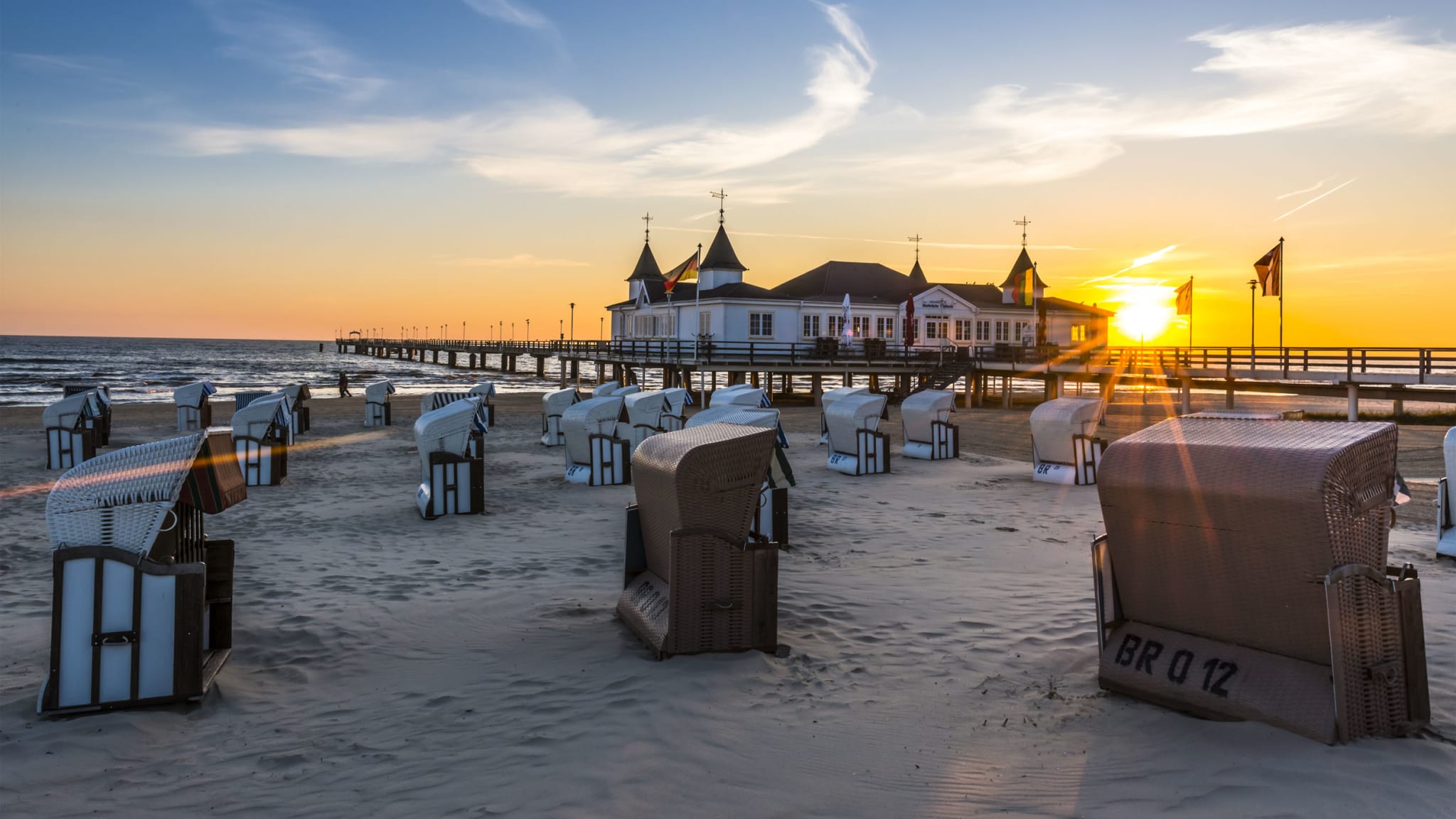Strandkörbe stehen an einem leeren Strand. Dahinter führt eine hölzerne Seebrücke aufs Meer hinaus.