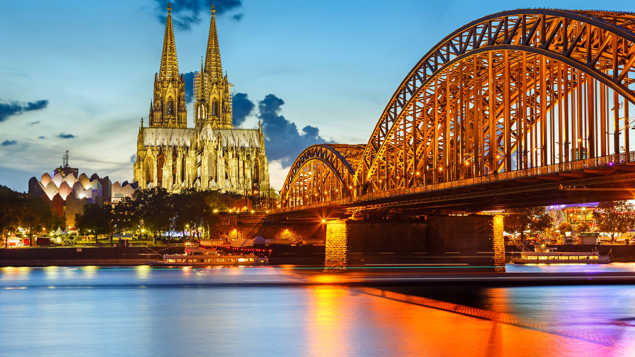 Blick über den Rhein auf die angeleuchtete Hohenzollernbrücke und den beleuchteten Kölner Dom im Abendlicht.