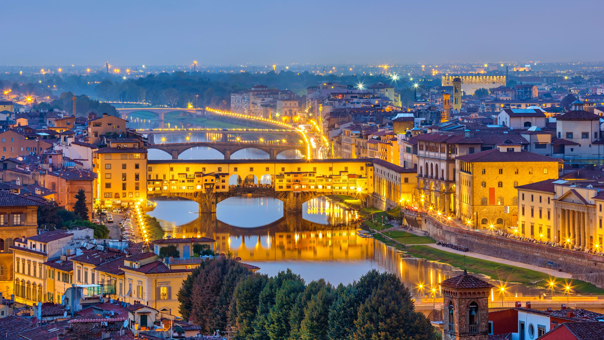 Blick auf die berühmte Ponte Vecchio bei Nacht. Die Brücke ist mit Häusern bebaut und hat in der Mitte große Rundbögen, durch die man auf den Fluss schauen kann.