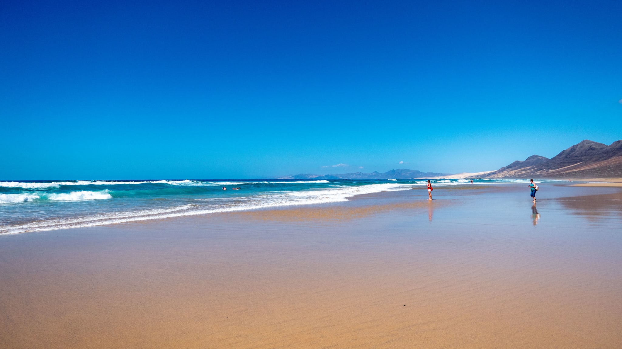 Ein weiter Strand mit tiefblauem, wolkenlosem Himmel und Bergen im Hintergrund.