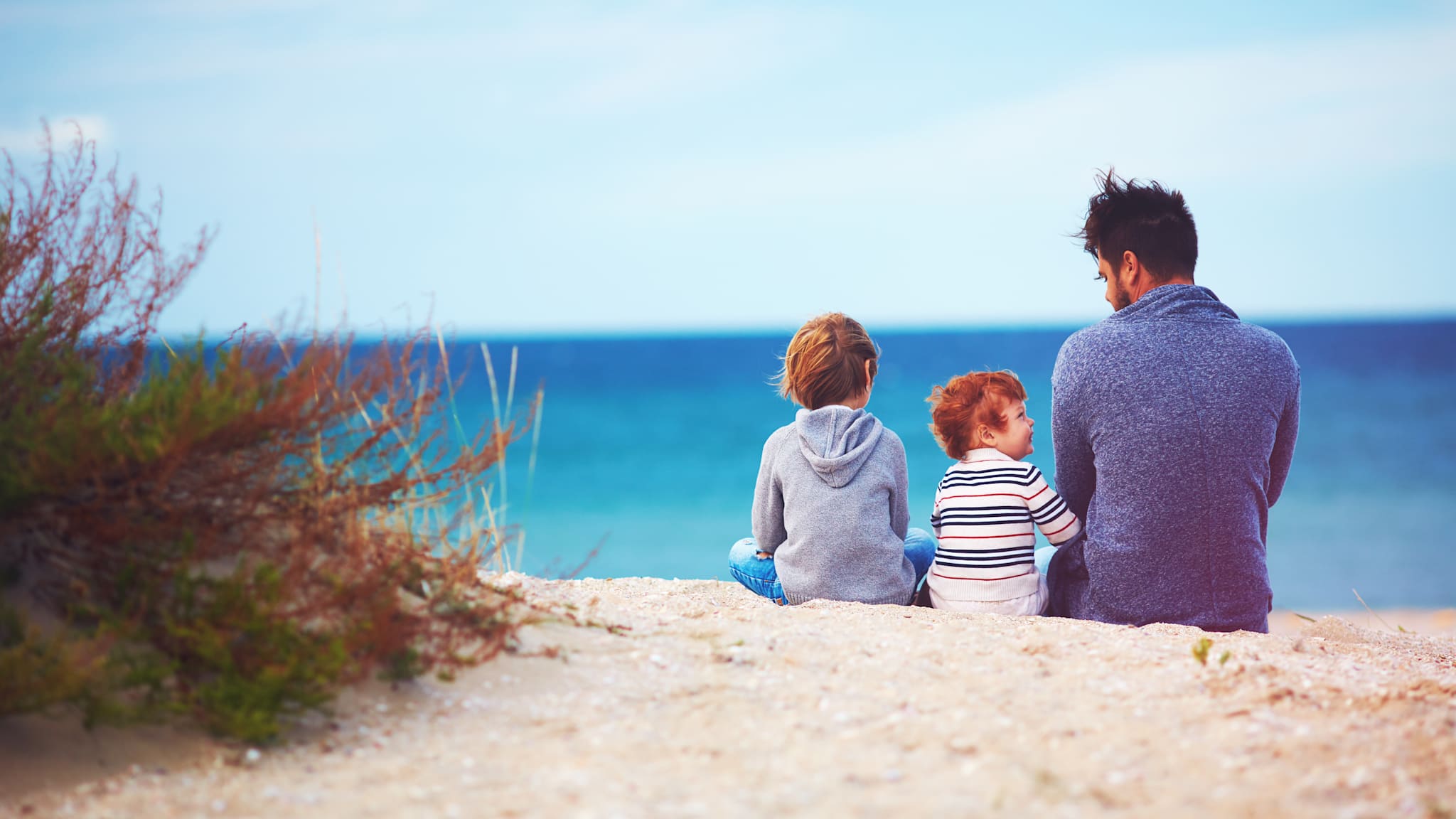 Vater sitzt mit seinen beiden kleinen Söhnen im Sand am Meer.