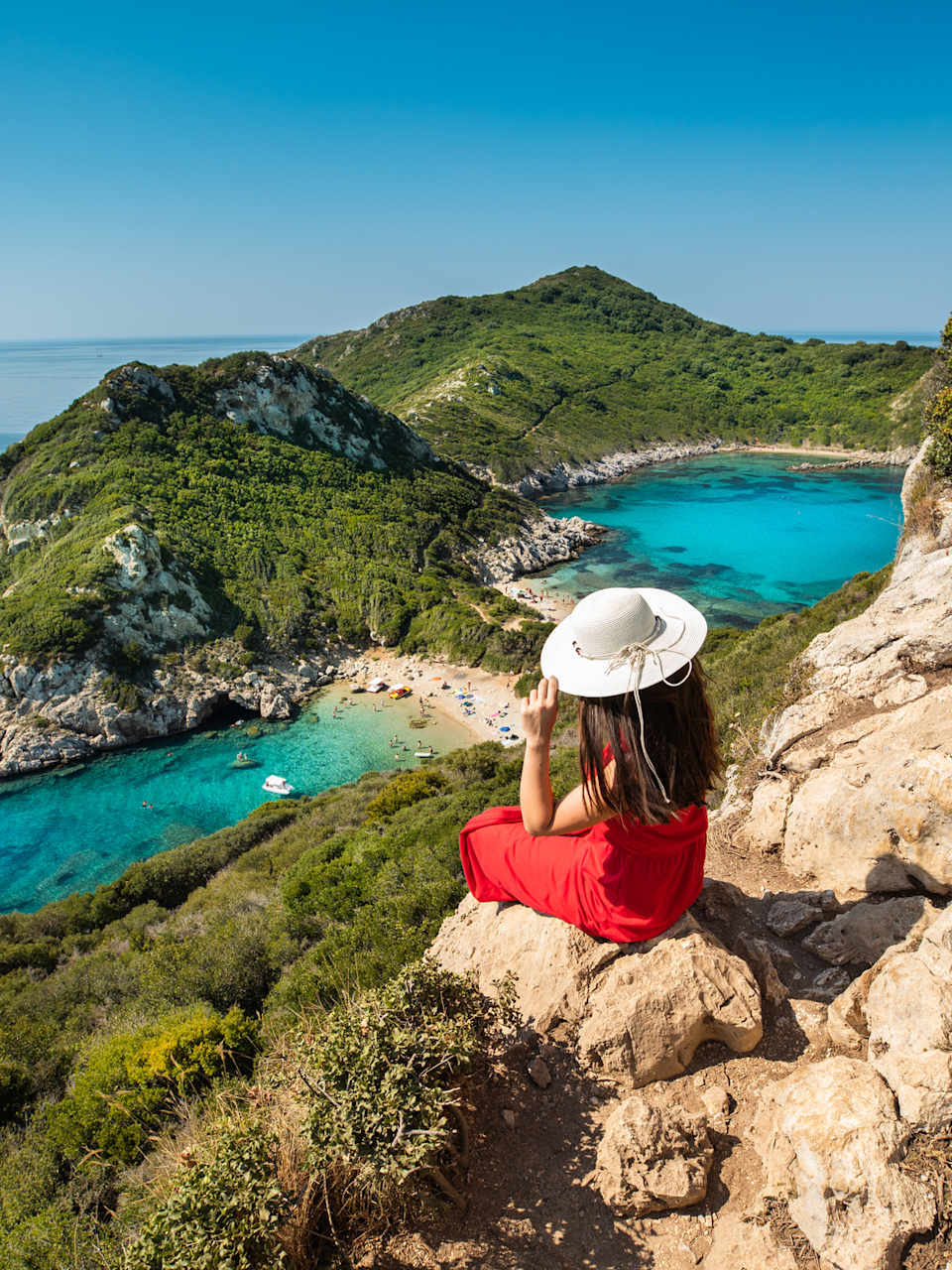 Frau mit Sonnenhut sitzt hoch oben auf einem Felsen und schaut auf Meer © iStock.com/RistoArnaudov