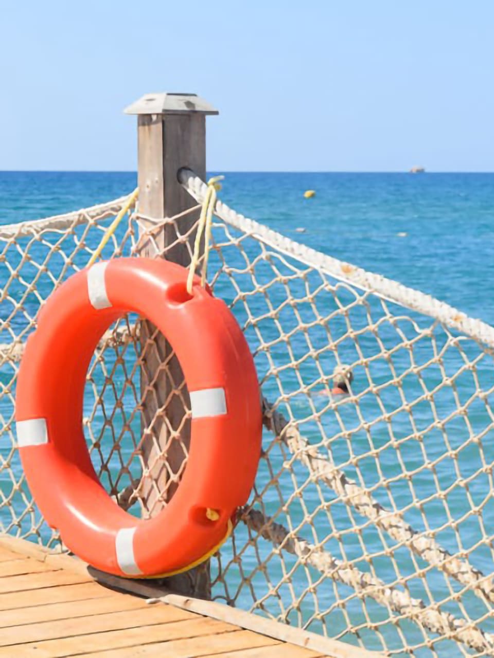 Blick von einem Steg aufs Meer. Am Steg hängt ein Rettungsring, während einige Personen im Meer schwimmen