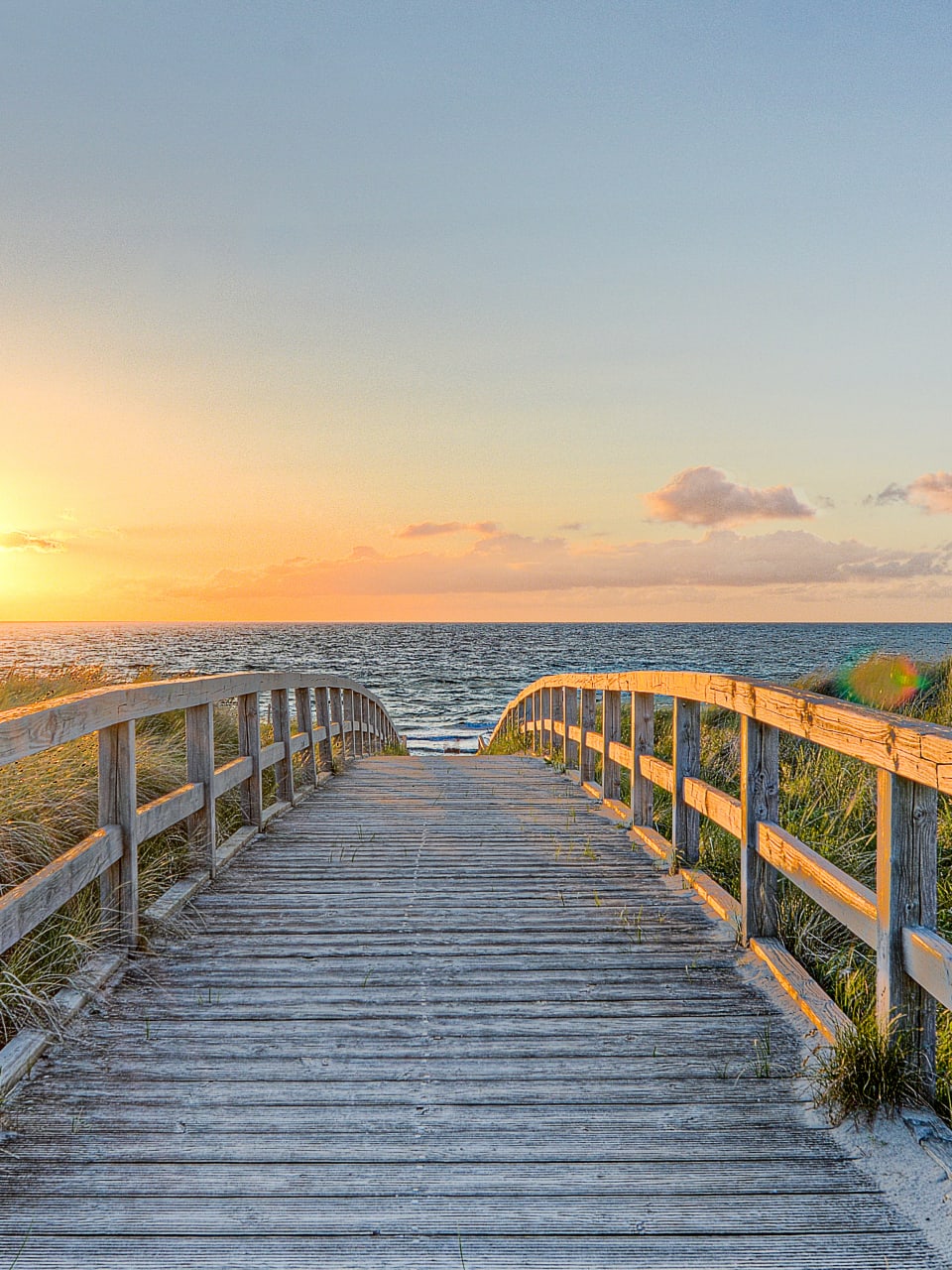 Ein langer Holzweg der Richtung Meer führt. Die Sonne geht unter und die Vögel fliegen über das Meer © Marco2811 - stock.adobe.com