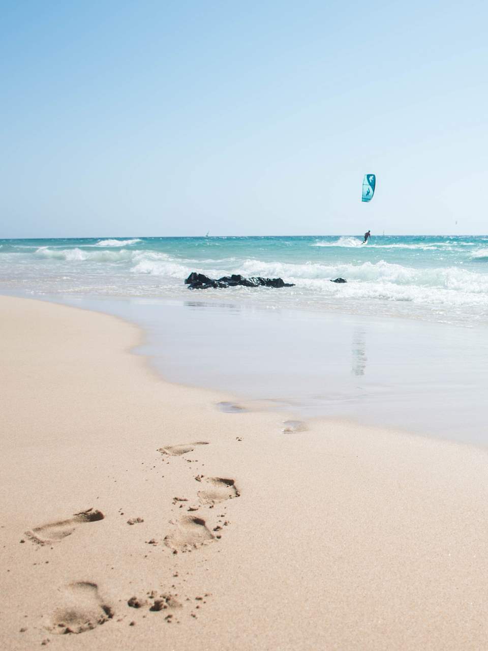 Sandstrand und blaues Meer mit Fußabdrücken und Menschen die kiteboarden © iStock.com/Pia Staric