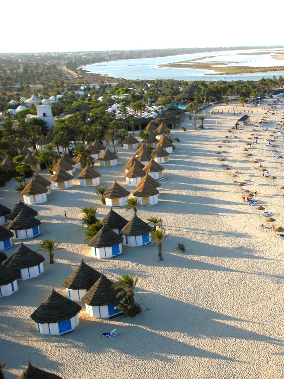 Drohnenaufnahme von einem hübschen Strand und dem blauen Meer. Auf dem Strand stehen einige Hütten, Sonnenschirme und Palmen.