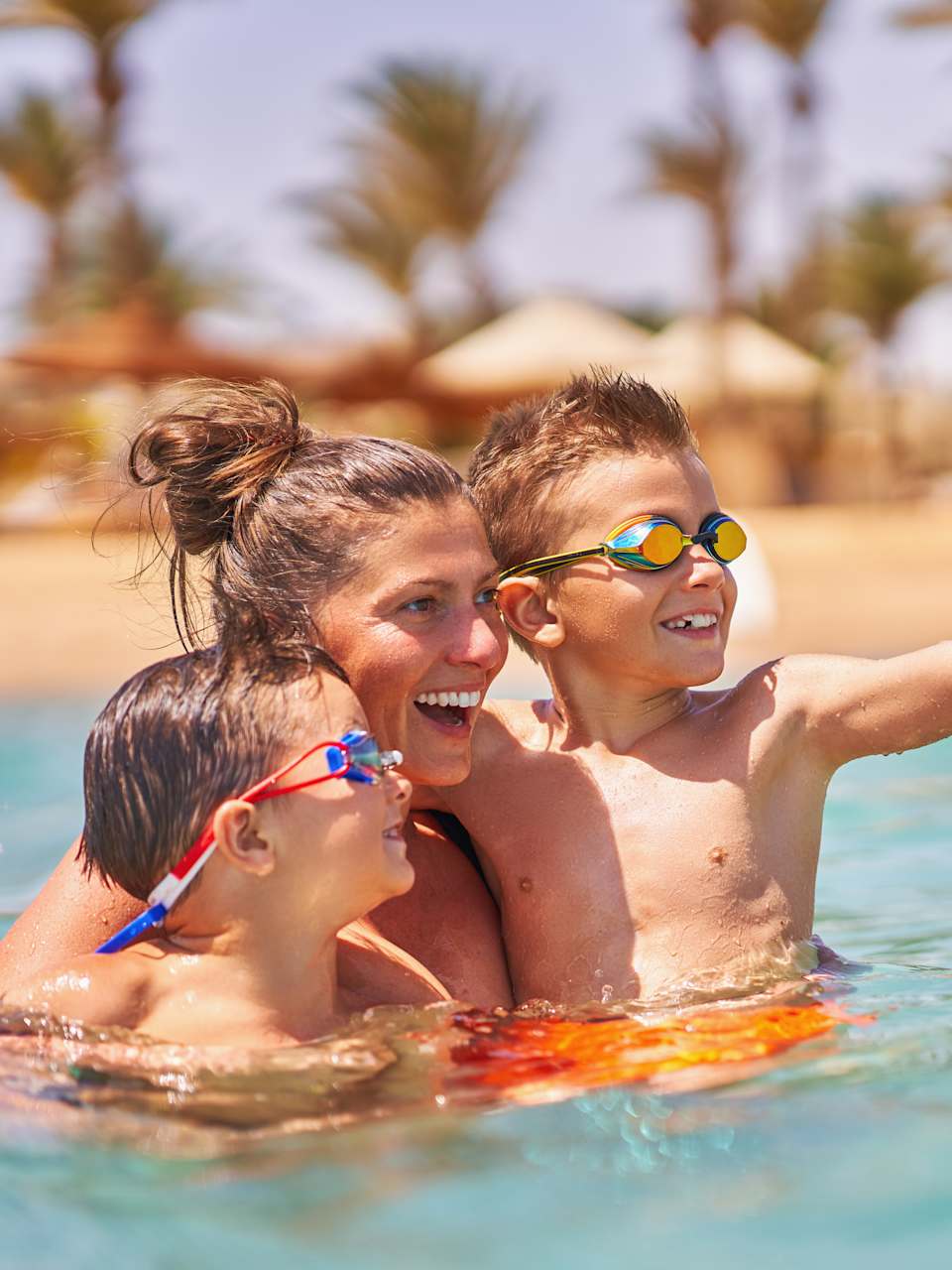 Eine Mutter ist mit ihren zwei Kindern im Wasser und hält sie im Arm. Im Hintergrund sieht man verschwommen den Strand und Palmen