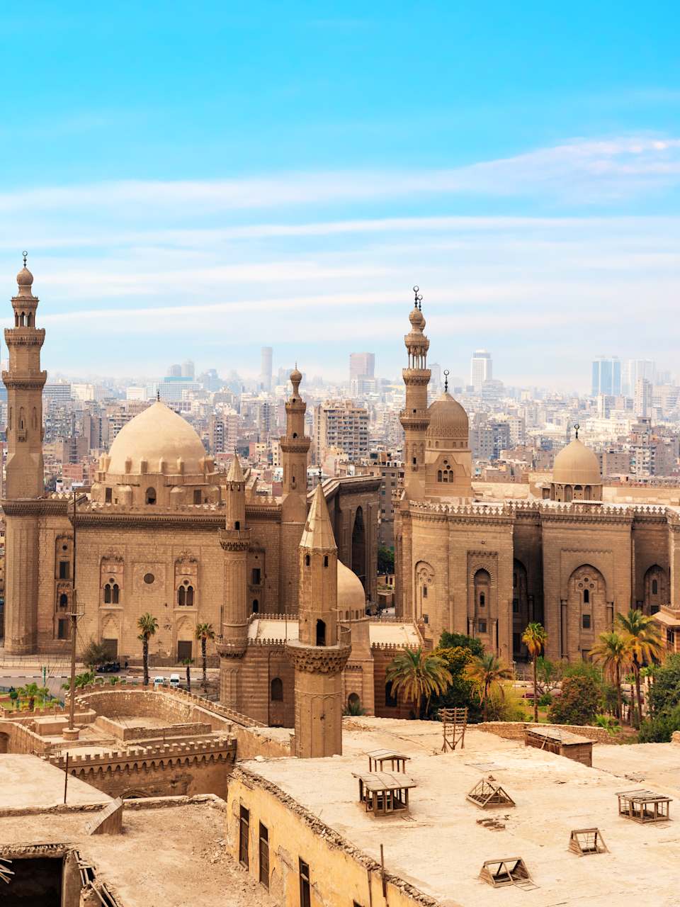 Die Moschee Madrassa ist im Vordergrund mit ihren vielen Türmen zu sehen. Im Hintergrund liegen unzählige Häuer der Stadt Kairo. In der Ferne erkennt man Pyramiden.
