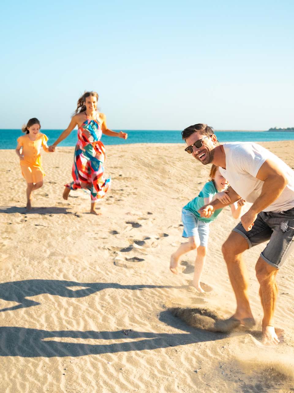 Eine Familie bestehend aus zwei Kindern, Mutter und Vater laufen spielerisch am Strand. Im Hintergrund ist das Meer zu sehen.