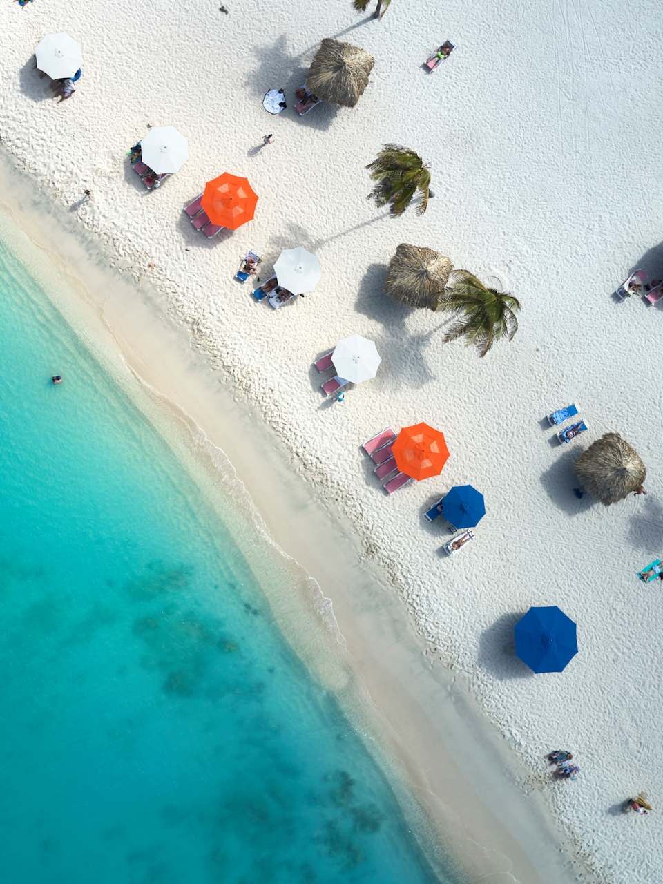 Ansicht von oben. Links ist das Meer, rechts ein Strand mit Sonnenschirmen, Palmen und Strandliegen.