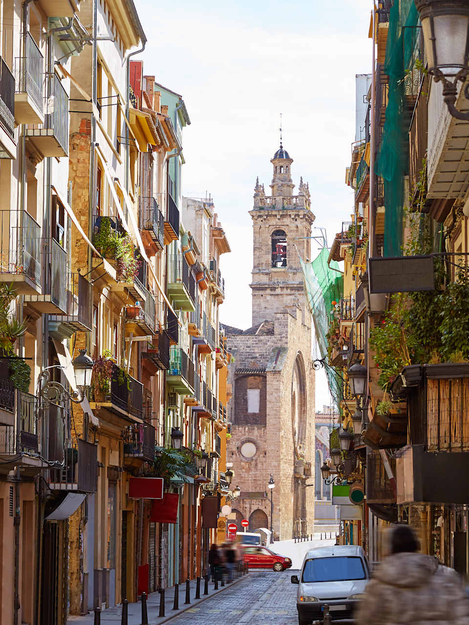 Blick durch eine schmale Straße in Valencia auf einen Platz mit einer großen Kirche. Die Häuser sind mit zahlreichen Balkonen bestückt, die hübsch mit Pflanzen dekoriert sind.