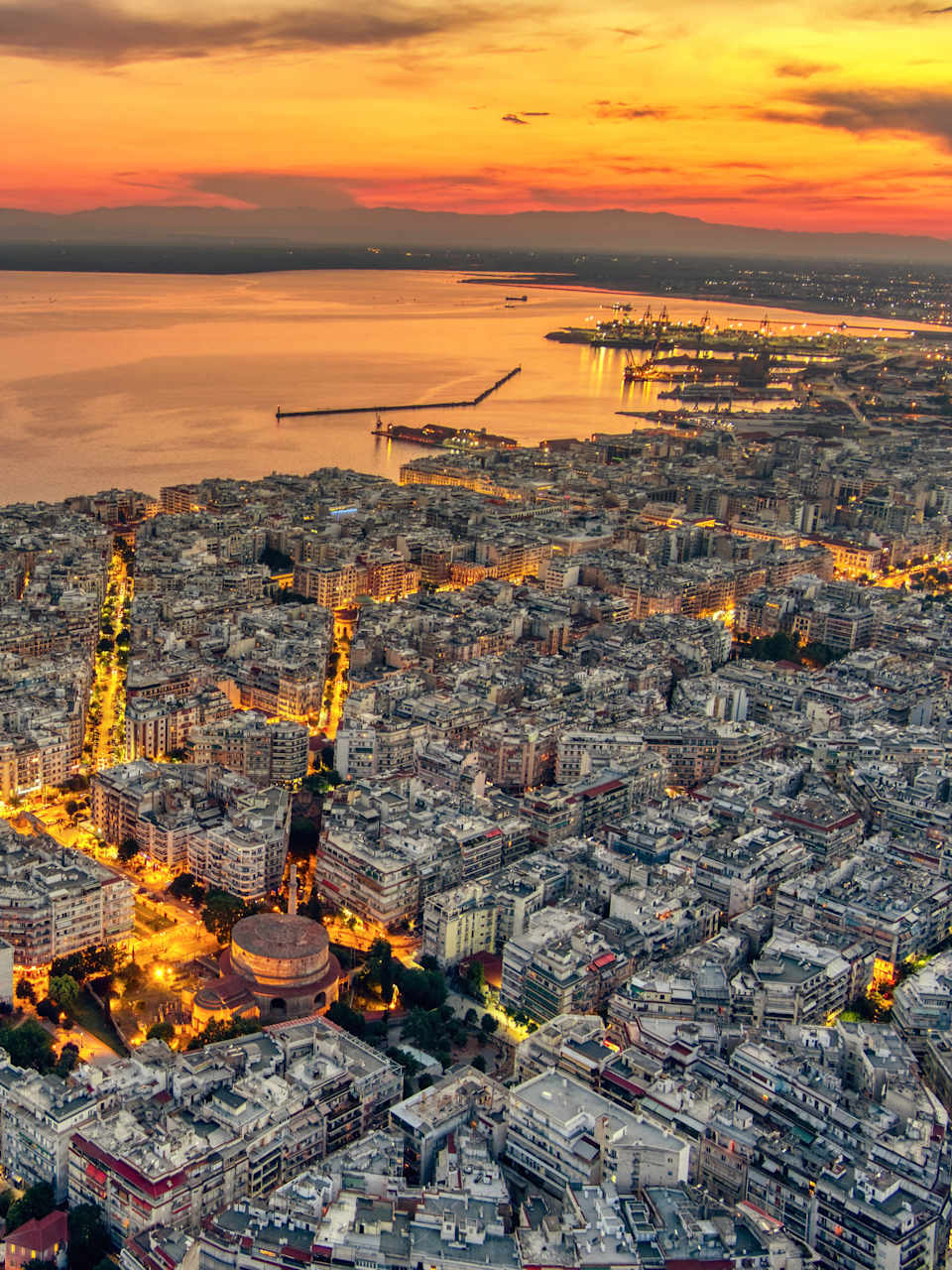 Vogelperspektive der Stadt Thessaloniki, Griechenland, bei Sonnenuntergang. Im Hintergrund ist das Meer. Die Straßen sind hell von den Lichtern der Stadt.
