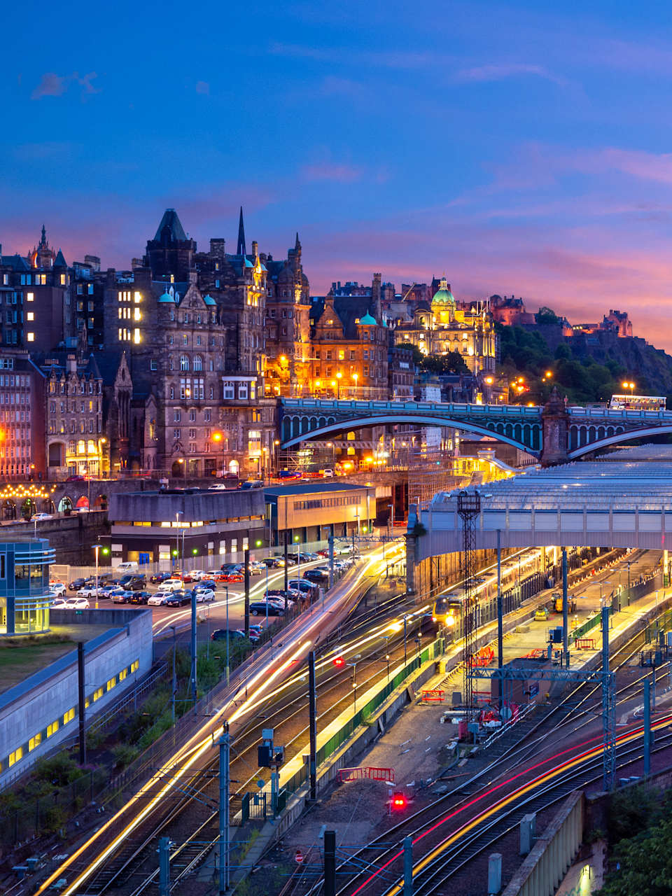 Die Gleise und das Bahnhofsgebäude in Edinburgh von oben. Dahinter erhebt sich ein kleiner Hügel, der mit  Häusern bebaut ist.