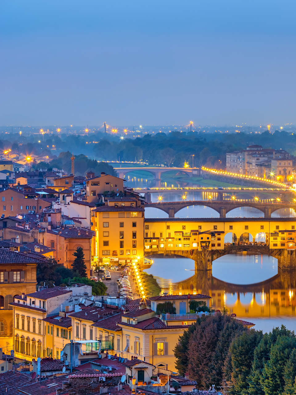 Blick auf die berühmte Ponte Vecchio bei Nacht. Die Brücke ist mit Häusern bebaut und hat in der Mitte große Rundbögen, durch die man auf den Fluss schauen kann.