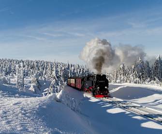 Eine schwarze Dampflok fährt durch eine tief verschneite Landschaft im Harz, umgeben von schneebedeckten Bäumen.