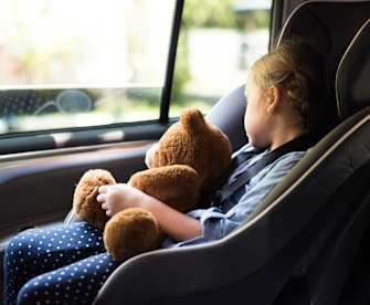 Ein Kind schläft im Kindersitz auf dem Rücksitz am Fenster mit einem Teddy im Arm.