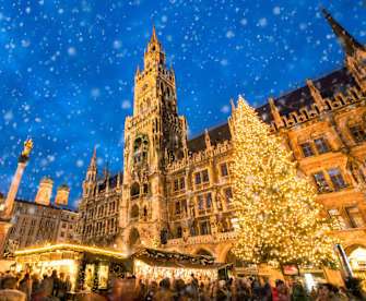 Winterliche abendliche Szene des Christkindlmarkts am Marienplatz in München, mit dem beleuchteten Rathaus, einem großen geschmückten Weihnachtsbaum und Schneefall über den Marktständen.
