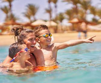 Eine Mutter ist mit ihren zwei Kindern im Wasser und hält sie im Arm. Im Hintergrund sieht man verschwommen den Strand und Palmen