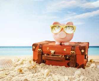 Sparschwein mit Sonnenbrille und Reisekoffer am Strand