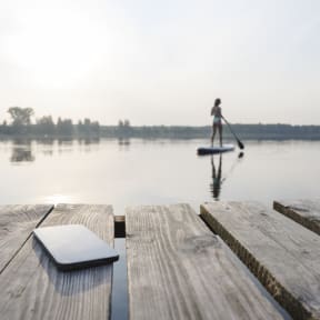Ein Handy liegt auf einem Holzsteg, während eine Frau auf dem See Stand-Up paddelt.
