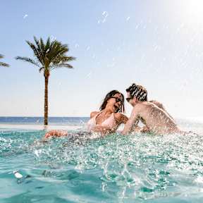 Ein Paar lacht im Pool mit Meer und einer Palme im Hintergrund.