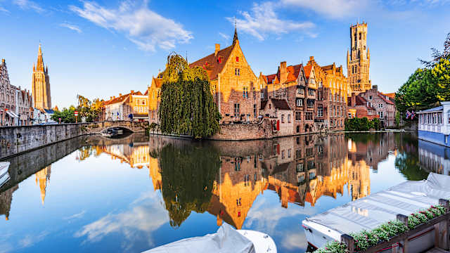 Ein mittelalterliches Bauensemlbe mit mehreren Häusern und einem Turm spiegelt sich im Wasser eines Kanals.