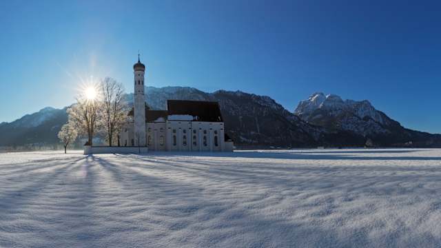 Panoramalandschaft in Bayern im Winter mit Wahrzeichen Kirche St. Koloman und Alpenbergen in der Nähe der Stadt Füssen, Bayern. © DieterMeyrl via Getty Images