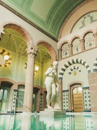 Eleganter Thermalraum mit großer Marmorstatue in einem Becken, umgeben von filigran bemalten Rundbögen, hohen Säulen, Jugendstil-Ornamentik und farbigen Wandfliesen im Palais Thermal Bad Wildbad.