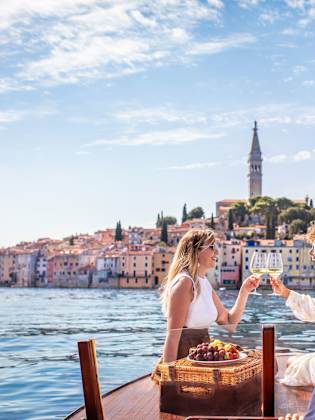 Zwei Frauen sitzen auf einem Boot auf dem Meer vor der Kulisse eines bunten Hafens in Kroatien und stoßen mit Weingläsern an
