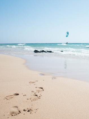 Sandstrand und blaues Meer mit Fußabdrücken und Menschen die kiteboarden © iStock.com/Pia Staric