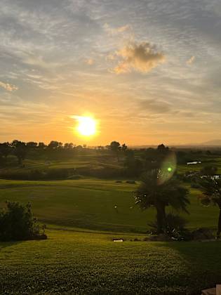 Ein weitläufiger, gepflegter Golfplatz mit Palmen und hügeligem Grün wird in warmes Licht eines Sonnenuntergangs getaucht, im Hintergrund Berge und weite Landschaft bei Palma de Mallorca.