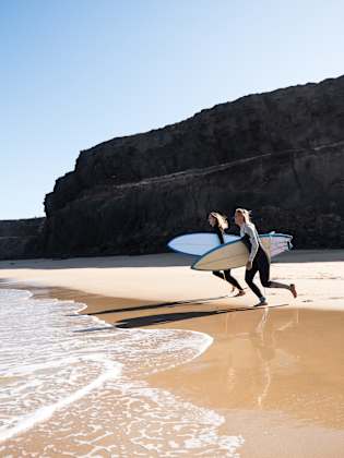 Surferinnen am Strandufer rennen zu den Wellen © iStock.com/1MEDIA