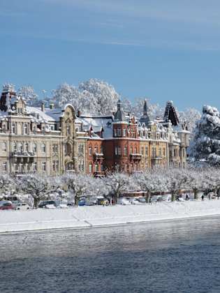 Historische Gebäude an der Uferpromenade von Konstanz, verschneite Bäume und ruhiges Wasser des Bodensees im Winter.