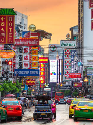 Autos fahren zwischen Hochhäusern entlang. Überall sind Werbeschilder mit thailändischer, chinesischer und englischer BEschriftung zu sehen.