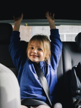 Ein kleines Mädchen sitzt fröhlich auf der Rückbank eines Autos. © Maskot/Maskot via Getty Images