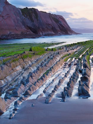 Man sieht moosbewachsene Steinrippen ins Wasser des Meeres ragen. Im Hintergrund sieht man eine große Klippe.