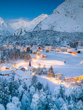 Alpendorf Maloja und Chiesa Bianca umrahmt von verschneiten Wäldern in der Abenddämmerung, Bergell, Kanton Graubünden, Engadin, Schweiz. © Roberto Moiola / Sysaworld via Getty Images