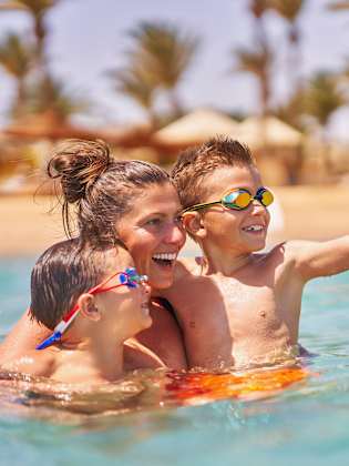 Eine Mutter ist mit ihren zwei Kindern im Wasser und hält sie im Arm. Im Hintergrund sieht man verschwommen den Strand und Palmen